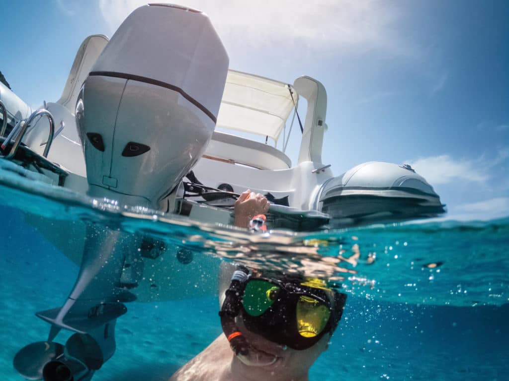 Half underwater selfie of a man with sea mask attached to the stern of an inflatable dinghy boat with outboard engine. The clean and clear sea of Villasimius in Sardinia.