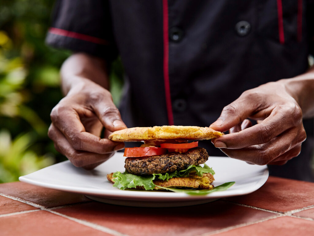 Chef Joachim Jammeal plating his mushroom burger with tomatoes, lettuce, vegan mayo and sandwiches between fried plantain cakes.