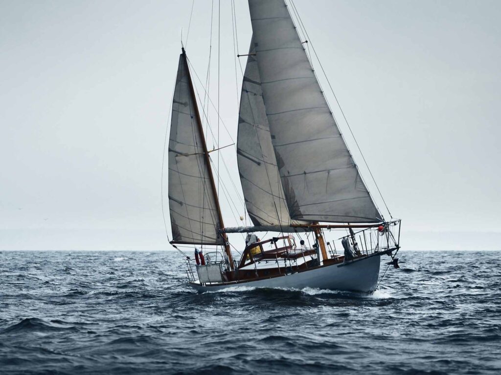 Old expensive vintage two-masted sailboat (yawl) sailing in an open sea during the storm. Reefed sails. Lighthouse in the background. Sport, regatta, racing, recreation, transportation. Panoramic view