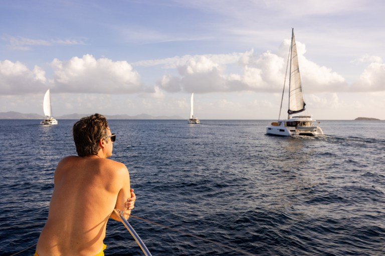 A man is sitting at the bow of a boat watching Moorings catamarans sail by.