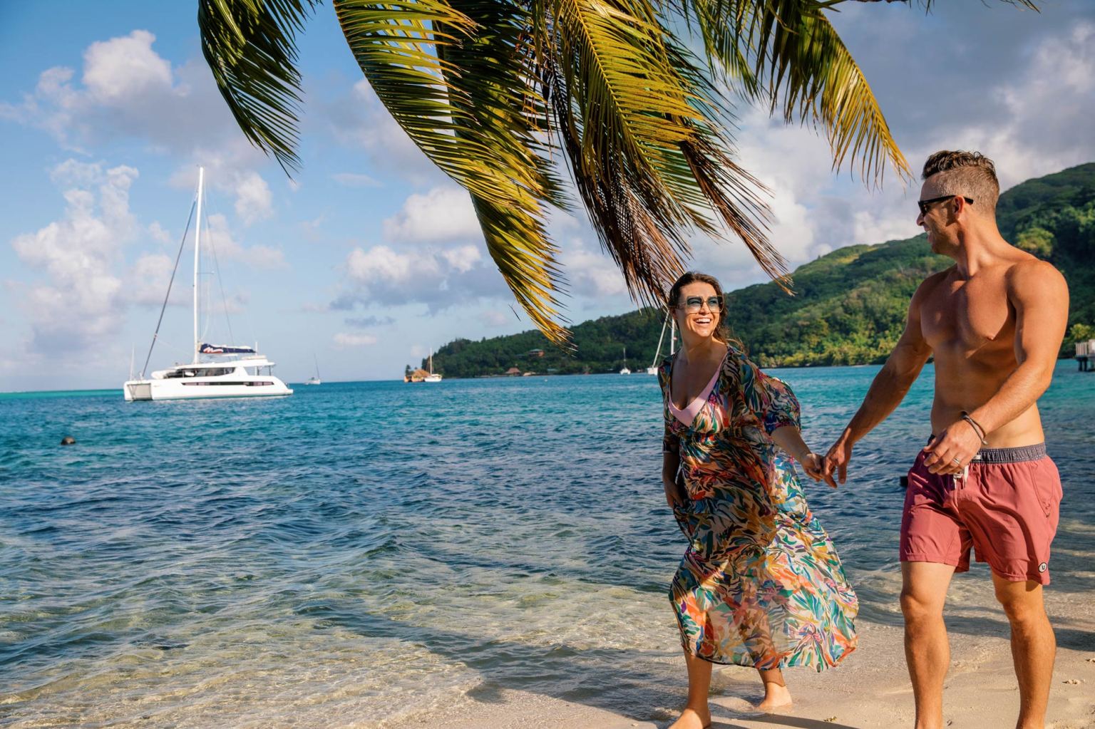 A couple walking along the beach in French Polynesia with a Moorings sailing catamaran in the background.