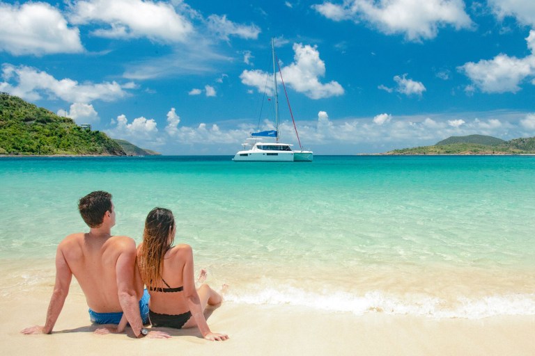 A couple sitting on the beach in the BVI with a Sunsail catamaran in the background.