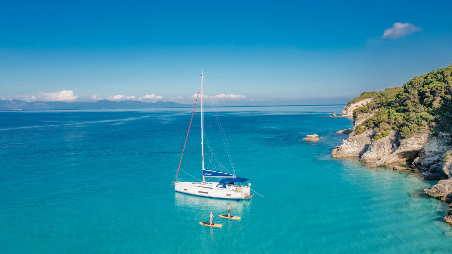 A Sunsail monohull at anchor in Corfu, Greece, with a couple paddleboarding nearby.